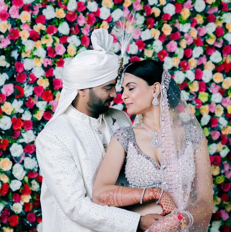 Groom in white sherwani and safa embracing the bride before a wall of roses