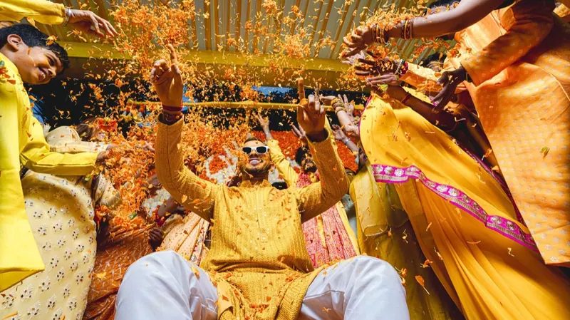 Groom in yellow kurta laughing as family throw marigold petals during the haldi
