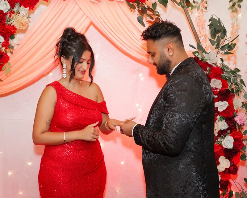 Man slips an engagement ring onto his partner's finger in front of a rose backdrop