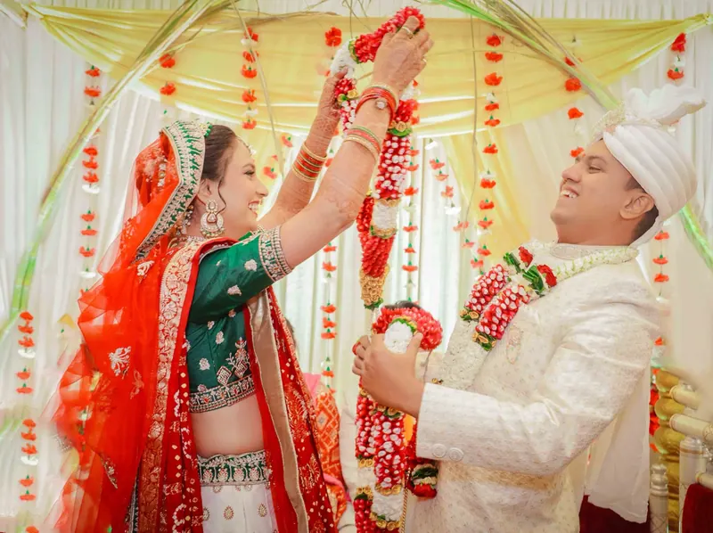 Bride lifts a rose and tuberose varmala over the groom under a marigold mandap
